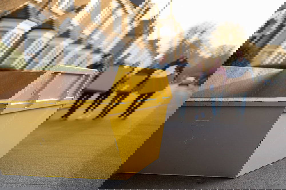 Multiple people walking towards a skip with boxes